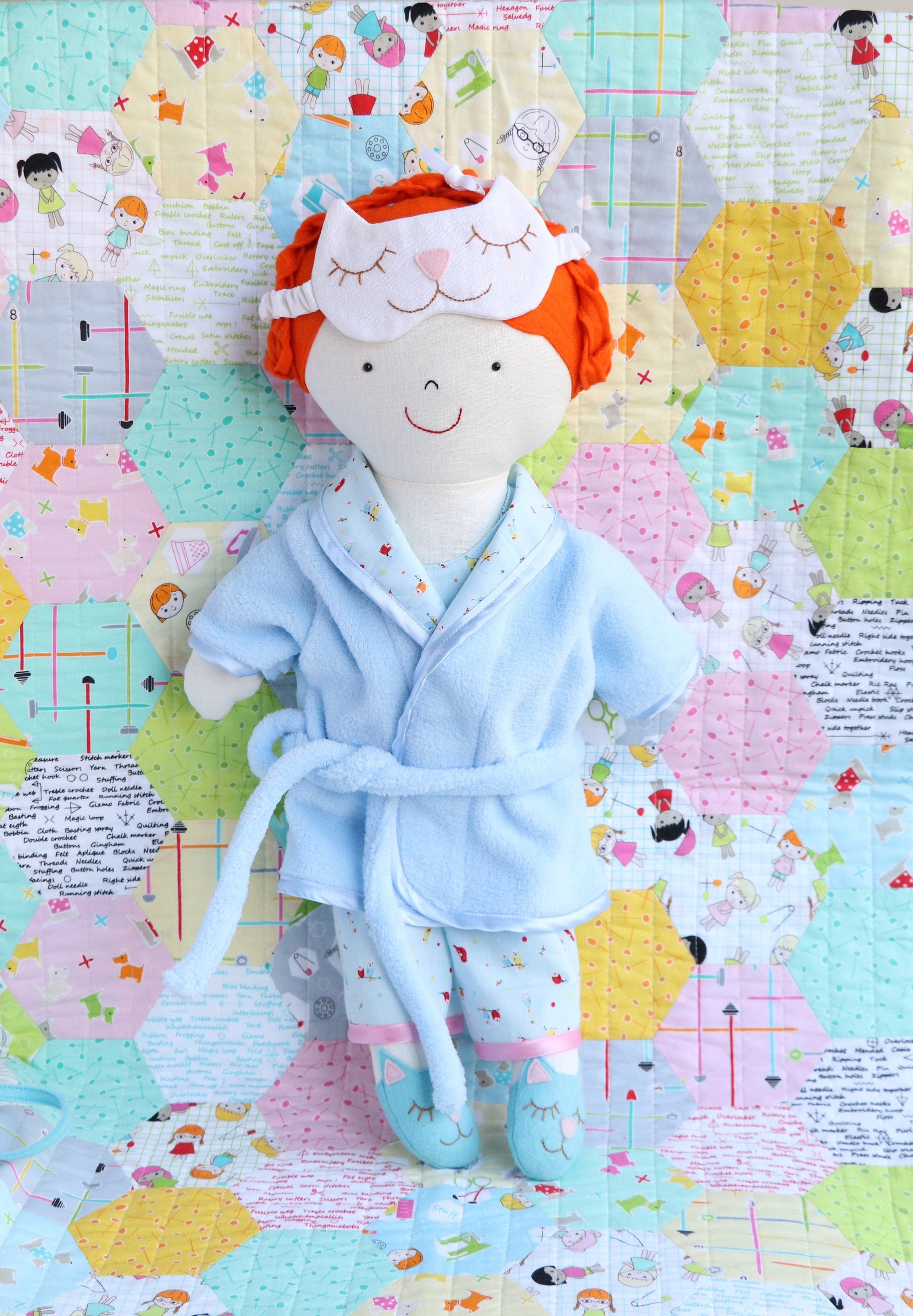 cloth doll with red hair wearing blue pyjamas and dressing gown. Doll is wearing a kitty shaped sleep mask
